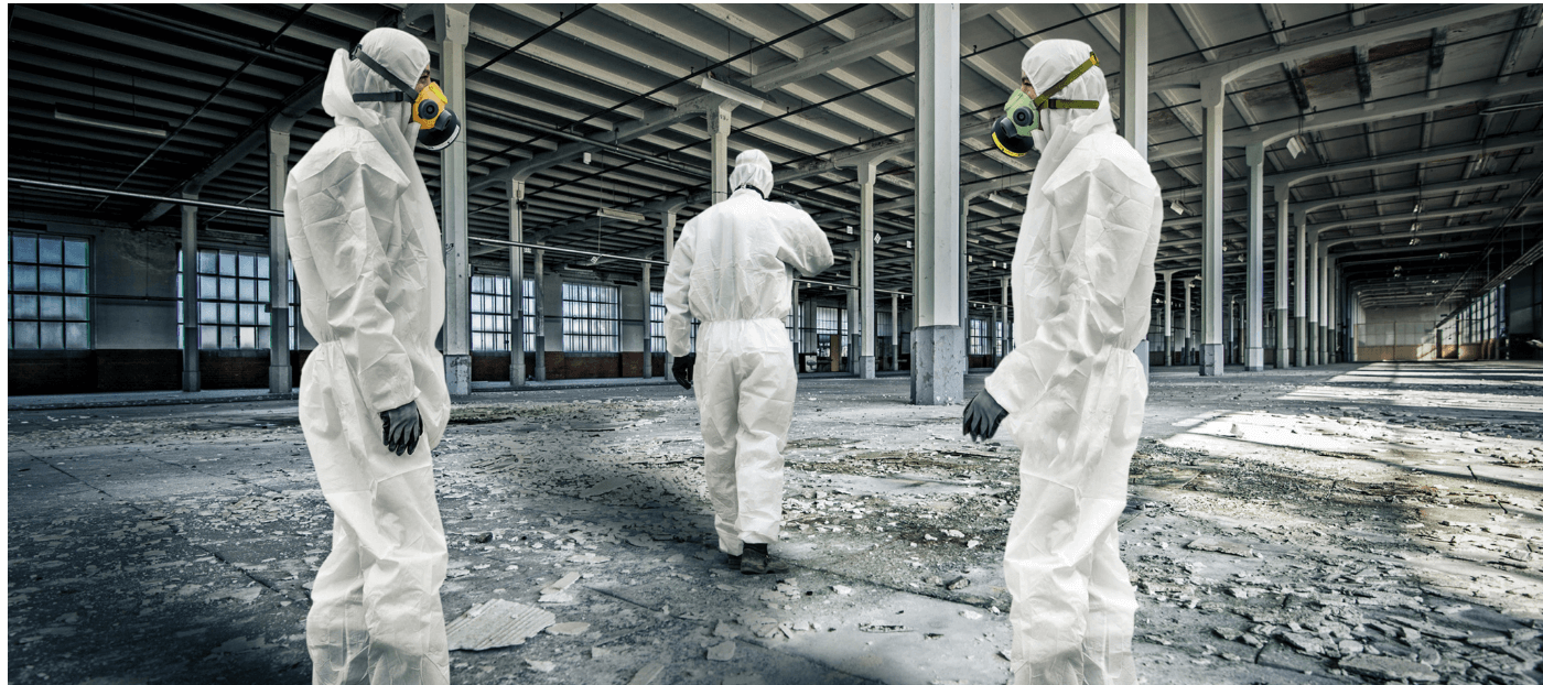 Drei Personen in weißer Schutzkleidung und Atemschutzmasken stehen in einer verlassenen, staubigen Halle mit großen Fenstern. Der Boden ist mit Schutt und Schmutz bedeckt, während die Personen sich in unterschiedlichen Positionen bewegen. Die Atmosphäre wirkt industriell und unbemannt.